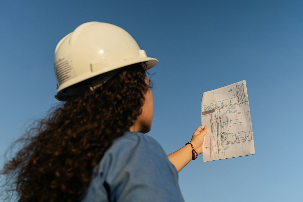 Iemand met een veiligheidshelm houdt een bouwplan omhoog tegen een blauwe lucht.